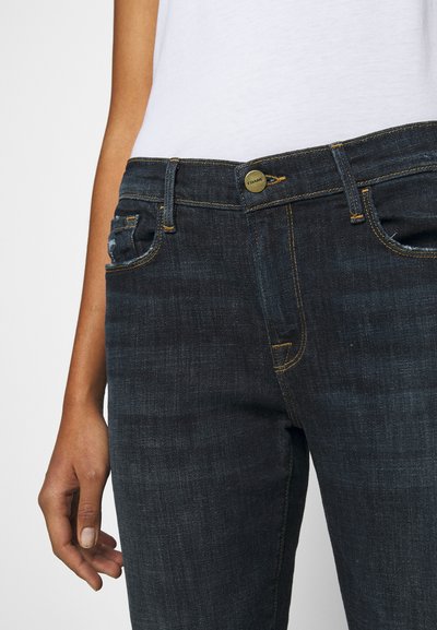 Jeans in denim blu scuro con un design aderente. Le caratteristiche includono una chiusura con bottone frontale, cuciture gialle e uno stile a cinque tasche.