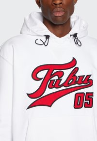 Felpa bianca con cappuccio caratterizzata da un grande logo rosso "Fubu" e il numero "05," con contorni neri e cordini regolabili sul cappuccio.