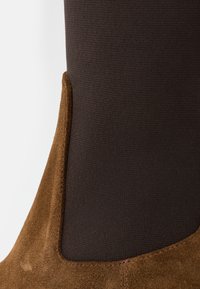 Bottine en daim marron avec tissu élastique foncé, présentant une texture lisse et des détails cousus le long du bord du daim.