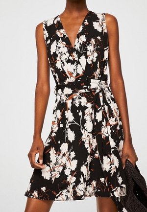 Robe noire sans manches avec imprimé floral blanc et orange, style portefeuille à col en V, ceinture nouée à la taille, portée par une personne debout.