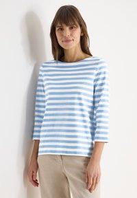 Femme aux cheveux bruns mi-longs portant une chemise à manches longues rayée bleu clair et blanc et un pantalon beige, debout contre un mur blanc.