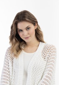 Witte opengewerkte cardigan met een losse structuur en een textuurpatroon, met een brede kraag en lange mouwen, gelaagd over een lichtgekleurde top.