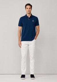 Polo shirt bleu marine avec col et manches courtes, associé à un pantalon blanc et des baskets foncées. Présente un petit logo brodé sur la poitrine.