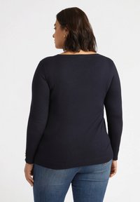 Granatowy sweter z długim rękawem i szerokim dekoltem, z rękawami i dolnym wykończeniem w prążki. W połączeniu z niebieskimi dżinsami, tkanina wydaje się być miękka i teksturowana.