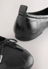 Paire de ballerines en cuir noir avec texture crocodile, avec un nœud sur une chaussure et une languette au talon sur l’autre.