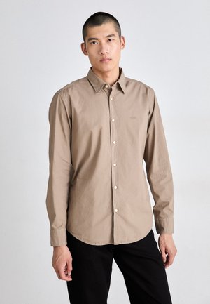 Light brown long-sleeve shirt with a button-down collar, featuring a subtle logo and cream buttons. Fabric has a smooth, soft texture.