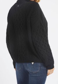 Schwarzer gestrickter Pullover mit strukturierten Zopfmustern, hohem Kragen und lockerer Passform. Getragen über blauen Jeans mit einem kleinen Markenetikett auf der Rückseite.