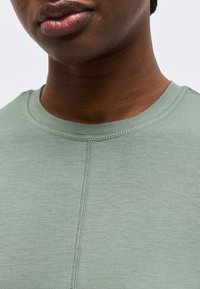 Κοντινό πλάνο ενός ατόμου που φοράει ανοιχτό πράσινο τοπ με στρογγυλή λαιμόκοψη και εμφανές ραφή που τρέχει κάθετα στο μπροστινό μέρος.