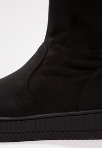 Primo piano di un stivale platform in suede nero che mostra la suola testurizzata e la cucitura dettagliata sul pannello laterale.
