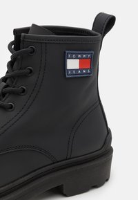 Černá kožená kotníková bota se silnou podrážkou, která má kovové okroužky, kulaté tkaničky a obdélníkovou nášivku s logem na boku.