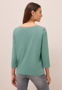 Femme aux longs cheveux bruns portant un haut à manches trois-quarts côtelé vert clair et un jean gris, vue de dos et légèrement tournée sur le côté.