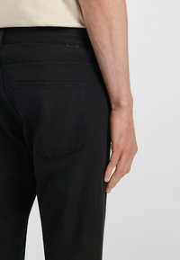 Black trousers made of cotton with a smooth texture, featuring subtle stitching, a back pocket, and a branded tag.