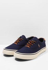 Námořnické plátěné tenisky s hnědými koženými akcenty, kulatá špička, šněrovací design, bílá gumová podrážka a logo na boku.