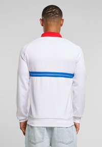 Felpa bianca con colletto rosso e striscia orizzontale blu. Presenta una texture liscia e polsini a coste sulle maniche e in vita.