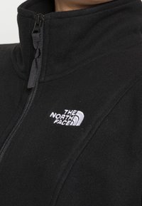 Musta fleecetakki, jossa on korkea kaulus, etumallinen vetoketju ja vasemmassa rintakehässä valkoinen kirjailtu logo.