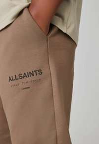 Ruskeat puuvillaiset joggerit, joissa on sileä pinta, ja sivussa musta printti "ALLSAINTS" sekä japaninkielisiä merkkejä.