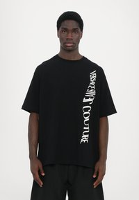 Svart oversized t-skjorte laget av bomull, med hvit vertikal tekst langs den ene siden. Rund hals og korte ermer, minimalistisk design.