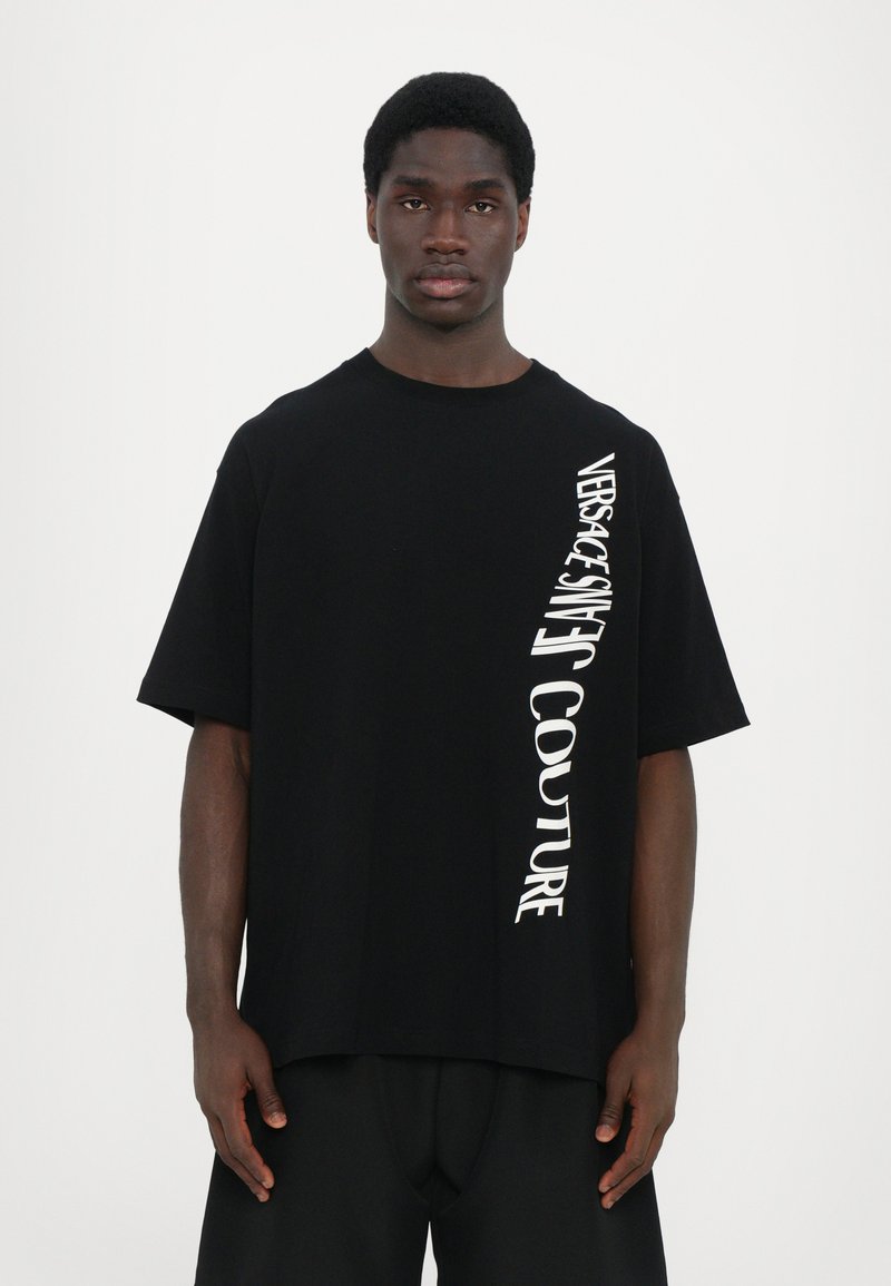 Svart oversized t-skjorte laget av bomull, med hvit vertikal tekst langs den ene siden. Rund hals og korte ermer, minimalistisk design.