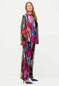 Completo a due pezzi colorato e patternizzato, composto da una giacca con bottone e pantaloni a gamba larga con forme audaci e righe in vari colori. Tacchi blu.