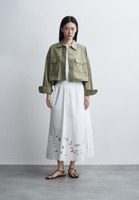 Femme debout contre un mur uni portant une veste courte olive, une jupe midi blanche avec des découpes florales et des sandales plates marron.