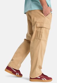 Persoon draagt beige cargobroek met zijzakken en rode Reebok sneakers, staand met één hand in de zak tegen een witte achtergrond.