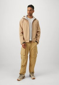 Beige vanntett jakke med svarte detaljer, grått fôr; beige cargobukser med flere lommer; grå skjorte; brune tursko med mønstrede lisser.