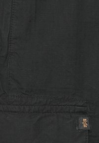 Tela negra con un patrón de cuadrícula texturizada, mostrando detalles de costura y una pequeña etiqueta de logo en la esquina inferior derecha.