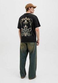 Must must juustega, millel on valge ja kuldne lilleline graafiline print seljal, kombineeritud vabade, kulunud roheliste denimite ja pruuni mütsiga.