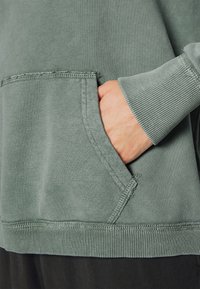 Mano con tono di pelle chiara dentro la tasca anteriore di una felpa verde sbiadita con polsini a costine e dettagli di cucitura visibili.