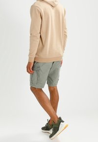 Felpa beige con cappuccio, pantaloni cargo verdi e sneaker verdi con dettagli bianchi. La felpa ha una texture morbida; i pantaloni presentano tasche laterali.