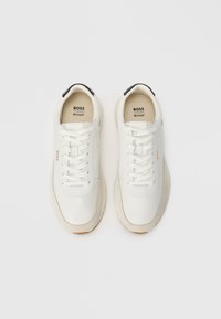 Hvite sneakers med en glatt læroverdel, tonede lisser og en lys brun såle. Svart aksent ved hælen, med "BOSS" merke.