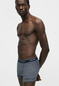 Άνδρας χωρίς πουκάμισο που φοράει μπλε και λευκά ριγέ boxers Calvin Klein, στέκεται με τα χέρια πίσω από την πλάτη σε απλό λευκό φόντο.