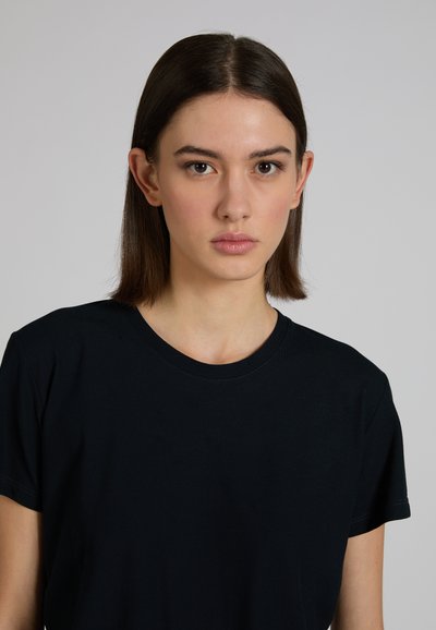 Jonge vrouw met stijlbruin haar, gekleed in een simpel zwart T-shirt, kijkt recht in de camera met een neutrale uitdrukking.