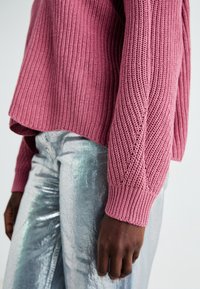 Pull rose tricoté avec une texture côtelée, design court, manches longues, associé à un pantalon argenté métallisé brillant.