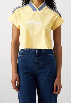 Femme portant un haut de sport Adidas court jaune avec des rayures bleues sur les épaules et un jean taille haute bleu foncé.