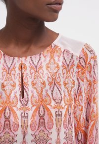 Flerfärgad blus med ett paisleymönster i rosa, orange och lila. Nyckelhålsutskärning med en mjuk, enfärgad axelpanel.