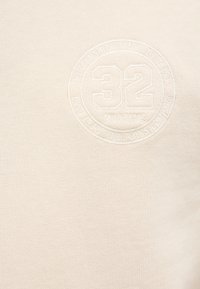 Kremowa bawełniana bluza z okrągłym, wypukłym logo z numerem "32" i napisem "MUSTANG". Teksturowana tkanina, minimalistyczny design.