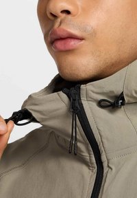 Beige jacka med hög krage, med svarta dragkedjor och snoddar, slät textur och synlig sömdetalj.