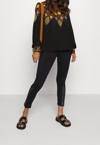 Maglietta nera con motivi geometrici colorati sul colletto e sui polsini, abbinata a leggings neri con dettagli testurizzati laterali e sandali neri.