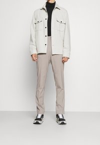 Lichtgrijze overhemden met twee borstzakken, zwarte coltrui eronder, beige broek en zwarte sneakers met witte zolen.