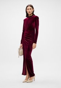 Μακρύ μανίκι φόρεμα από βελούδο μπουρνιώτικου χρώματος με  gathered λεπτομέρειες και πλαινές σχισμές, σε συνδυασμό με μια μπεζ τσάντα από καστόρι και ροζ κουνάδι. Λείο ύφασμα.