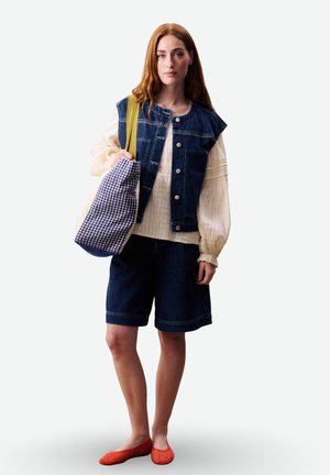 Giovane donna che indossa un gilet di jeans e pantaloncini, una blusa color crema, ballerine rosse, con una borsa a tracolla a quadretti con una cinghia gialla sulla spalla.
