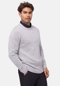 Grauer Rundhalsausschnitt-Pullover aus weichem Strickmaterial, mit gerippten Ärmelabschlüssen und Saum. Wird über einem navyblauen Hemd mit Kragen getragen, die Hände in den Taschen.
