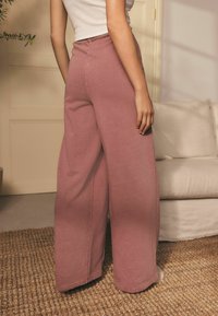 Bredde ben, lyserøde sweatpants lavet af bl�ødt stof, med elastisk talje og minimale syninger. Baggrunden har møbler i neutrale farver.