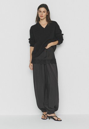 Femeie cu păr lung și închis la culoare, purtând un pulover negru larg cu guler în V, pantaloni negri largi cu dantelă și sandale negre cu vârf deschis, stând lângă un perete alb.