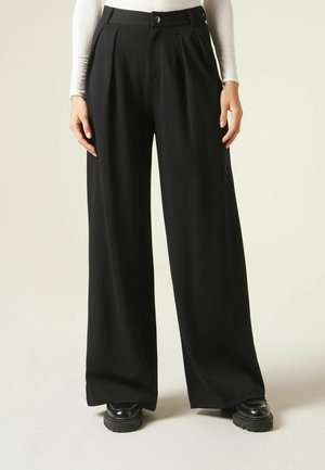 Femme portant un pantalon noir taille haute à jambes larges avec plis devant et fermeture par boutons, associé à des chaussures noires épaisses et un haut blanc à manches longues.