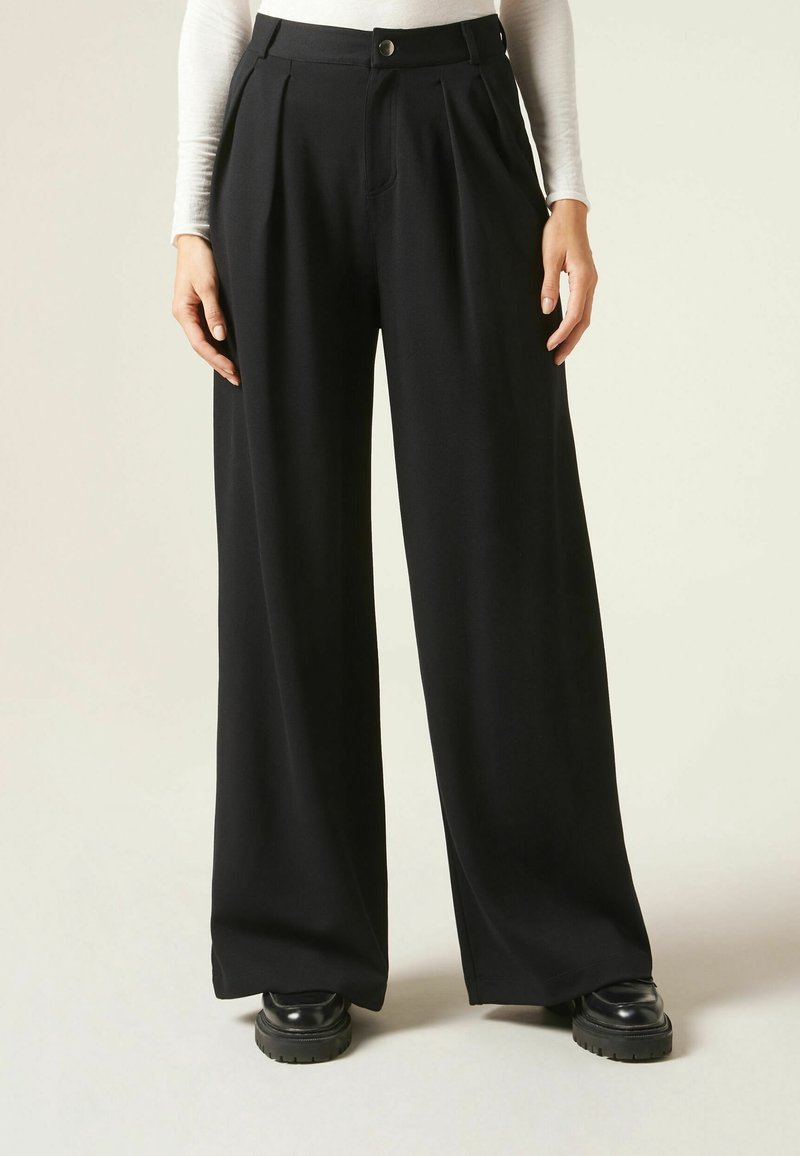 Femme portant un pantalon noir taille haute à jambes larges avec plis devant et fermeture par boutons, associé à des chaussures noires épaisses et un haut blanc à manches longues.