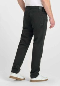 Man in zwarte rechttoe rechtaan broek en witte sneakers, staande tegen een effen lichte achtergrond, gezien vanaf de achterkant.