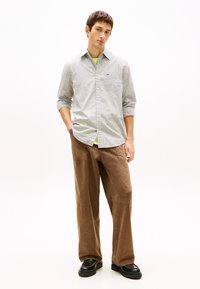 Νέος άνδρας που φοράει ανοιχτό καρό πουκάμισο με τα μανίκια γυρισμένα, καφέ παντελόνι φαρδύ στο πόδι και μαύρα παπούτσια, στέκεται με το ένα χέρι στην τσέπη.
