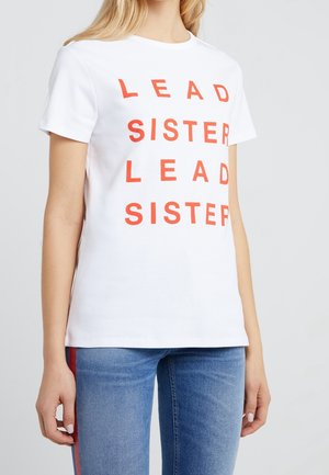 Vrouw draagt een wit T-shirt met rode tekst "LEAD SISTER LEAD SISTER" gecombineerd met een blauwe spijkerbroek met rode zijstrepen.